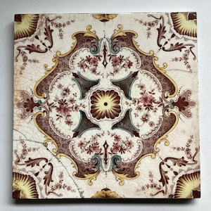 May include: Square ceramic tile with a floral pattern. The tile has a central flower design, framed by a brown and gold ornate border. The background is off-white with cracks, and the flowers are pink and burgundy.