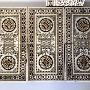 May include: A set of rectangular ceramic tiles featuring a symmetrical pattern in shades of cream, brown, and black. Each tile displays two circular floral motifs, geometric borders, and stylised butterfly silhouettes along the outer edges.