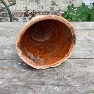 May include: A weathered terracotta plant pot, viewed from above. The pot has a wide opening and tapers inwards. The interior shows signs of age and use, with dark patches and a rough texture. The rim is slightly chipped.