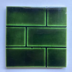 May include: A square ceramic tile featuring a glossy, deep forest green glaze with a subtle crackle finish. The surface is divided into a brick-like pattern by dark, recessed grout lines, creating a structured, rectangular grid arrangement.