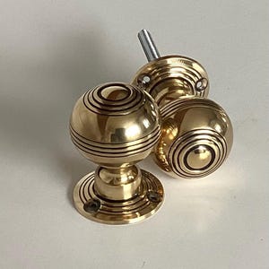May include: Three brass door knobs with a spherical design. Each knob features concentric circular grooves and a round base with screw holes. The knobs have a shiny, golden finish, suggesting a vintage or antique style.