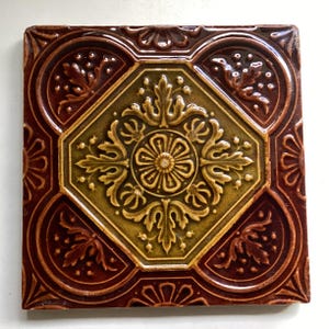 May include: Square decorative tile with an embossed floral design. The tile features a central octagonal shape in olive green, surrounded by a detailed, golden floral pattern. The outer border is a deep reddish-brown with additional floral motifs.