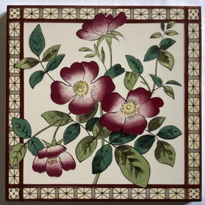 May include: A vintage tile featuring a floral design with pink roses and green leaves on a white background. The tile has a brown border with a geometric pattern.