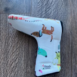May include: A white golf club head cover with a colourful dog pattern. The cover features various dog illustrations in brown, green, and blue. The text "4 Heads handmade" is visible on the cover.