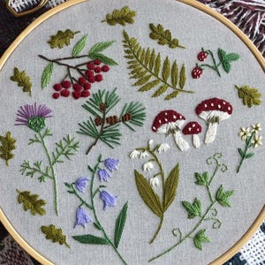 May include: An embroidered artwork in a wooden hoop, showcasing a botanical theme. The design includes red berries, mushrooms, ferns, and wildflowers in green, red, white, and purple. A pair of gold scissors is placed near the hoop, adding a touch of craftsmanship.