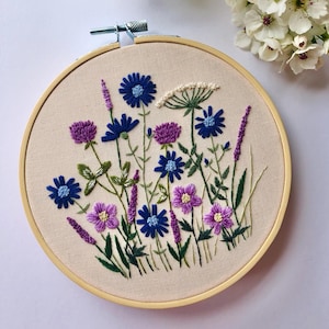 May include: Embroidered hoop art featuring a variety of purple and blue flowers with green stems and leaves on a beige fabric background.