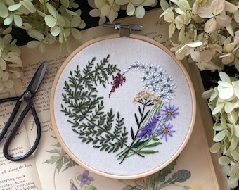 May include: A close-up of a circular embroidery hoop with a floral design. The embroidery features a variety of flowers in shades of purple, white, and green, including ferns, primroses, and forget-me-nots. The hoop is set against a background of a book with botanical illustrations.