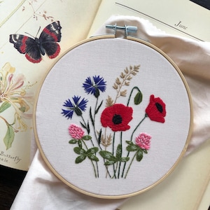 May include: An embroidered floral design in a wooden hoop. The design features red poppies, blue cornflowers, and pink clover, with green leaves and wheat stalks. The hoop is set against an open book with a butterfly illustration.