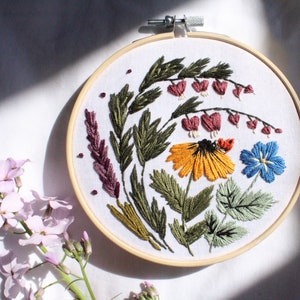 May include: Embroidered hoop art featuring a variety of flowers and greenery in shades of green, yellow, pink, blue, and red. The flowers are arranged in a circular pattern and are surrounded by leaves.