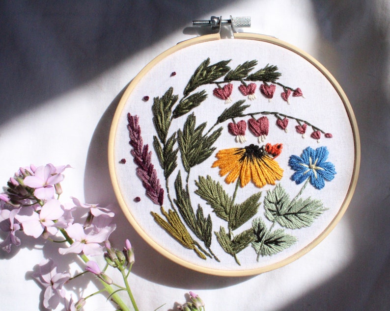 May include: Embroidered hoop art featuring a variety of flowers and greenery in shades of green, yellow, pink, blue, and red. The flowers are arranged in a circular pattern and are surrounded by leaves.