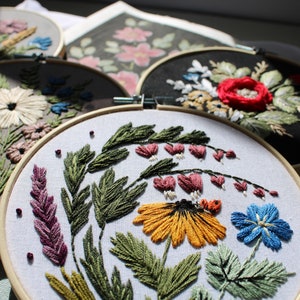 May include: Four embroidery hoops featuring floral designs. The hoops are made of wood and have white fabric stretched over them. The designs include a variety of flowers, leaves, and stems in shades of pink, red, yellow, blue, and green.