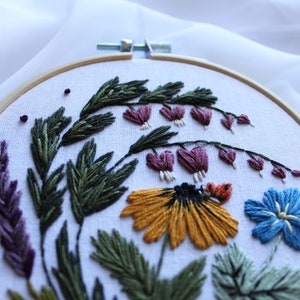 May include: Embroidered hoop art featuring a variety of flowers, including a yellow daisy, a blue flower, and purple bleeding hearts. The flowers are surrounded by green leaves and stems.