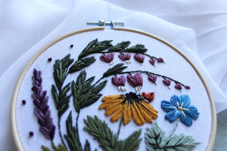 May include: Embroidered hoop art featuring a variety of flowers, including a yellow daisy, a blue flower, and purple bleeding hearts. The flowers are surrounded by green leaves and stems.