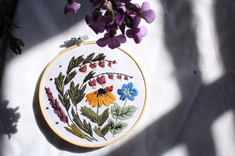 May include: Embroidered hoop art featuring a variety of flowers in shades of pink, yellow, blue, and green. The flowers are arranged in a circular pattern on a white background.