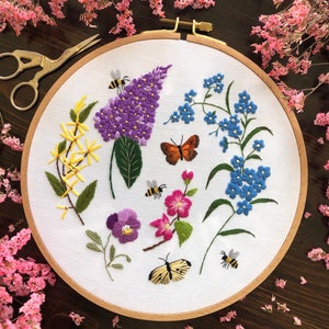 Könnte beinhalten: Stickerei-Bild in einem Reifen mit verschiedenen Blumen, darunter lila Flieder, blaue Vergissmeinnicht, gelbe Blüten, rosa Blüten und eine violette Stiefmütterchen. Es sind auch zwei Schmetterlinge und drei Bienen auf dem weißen Stoff gestickt.