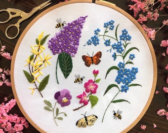 May include: Embroidered hoop art featuring a variety of flowers, including purple lilac, blue forget-me-nots, yellow blossoms, pink flowers, and a purple pansy.  There are also two butterflies and three bees embroidered on the white fabric.