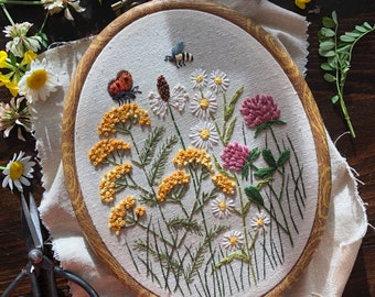 May include: Embroidered hoop art featuring a variety of wildflowers, including yellow yarrow, white daisies, and a pink clover. The hoop is made of wood and has a natural finish.
