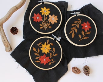 May include: Three embroidery hoops with black fabric and colorful floral embroidery designs. The hoops are on a black fabric background.