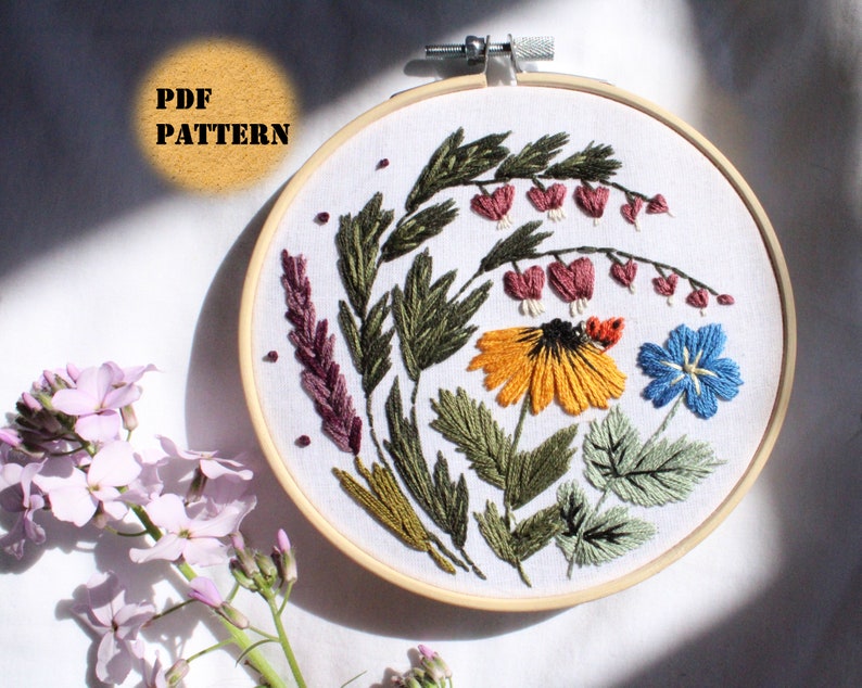 May include: Embroidered hoop art featuring a floral design with a variety of flowers, including a yellow flower with a black centre, a blue flower, and pink flowers. The flowers are surrounded by green leaves and stems.