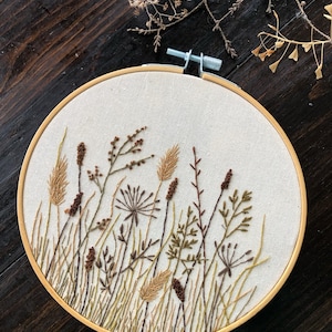 Op de afbeelding: Een ronde borduurring met een witte stoffen achtergrond met een gedetailleerd borduurmotief van verschillende bruine en groene grassen en planten.