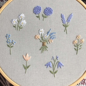 May include: An embroidery hoop with a grey fabric background, showcasing embroidered flowers in blue, white, and peach hues. The floral designs include daisies and lavender, with green stems. The hoop is made of wood.