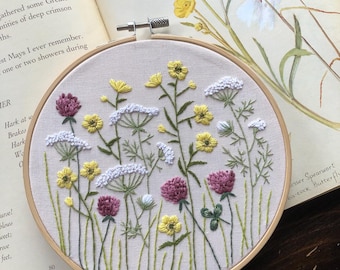 May include: An embroidered floral hoop art piece featuring a field of wildflowers in yellow, white, and purple. The embroidery is set against a beige fabric, framed by a wooden hoop. A butterfly illustration is visible in the background.
