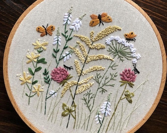 May include: Embroidered hoop art featuring a floral design with pink, white, and yellow flowers, green leaves, and orange butterflies. The hoop is made of wood and has a beige fabric background.