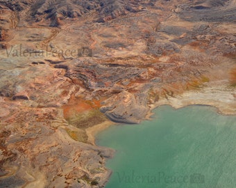 May include: An aerial view of a desert landscape with a turquoise lake. The land is a mix of brown, orange, and white, with a few green patches of vegetation. The lake is surrounded by rocky cliffs.