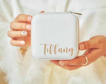 May include: A white, square-shaped jewelry box with a zipper closure. The box has the name "Tiffany" written in gold glitter lettering.