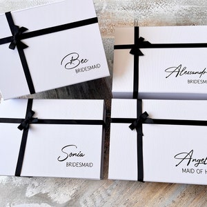 Puede incluir: Cuatro cajas de regalo blancas con cinta negra y lazos. Cada caja tiene un nombre escrito en cursiva en la parte delantera, con el título "BRIDESMAID" o "MAID OF HONOR". Los nombres son: Bec, Alessandra, Sonia y Angela.