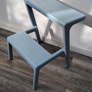 Op de afbeelding: Een lichtblauwe opstapje met twee treden en een gladde, matte afwerking. Het opstapje is voorzien van een lagere trede en een hoger bovenplatform, ondersteund door vier stevige poten. Het ontwerp is eenvoudig en functioneel met afgeronde hoeken.