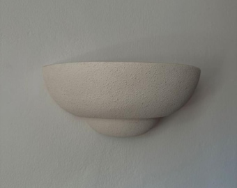 May include: A semi-circular wall sconce featuring a textured, off-white finish. The fixture has a bowl-shaped upper section that tapers down to a smaller, rounded base, creating a layered, stepped profile against the wall surface.