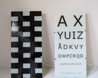 Pode incluir: Dois painéis retangulares de vidro. O painel esquerdo apresenta um padrão xadrez em preto e branco. O painel direito é uma tabela de exame de vista branca com letras pretas em tamanhos decrescentes: A X, YUIZ, ÅDKVY, DWPCRQD, ANMJHGDSIYJA e DOCFGDÅFCXKFYCJNW.