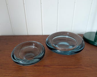 May include: Two circular glass bowls with a thick, rounded rim and a deep, concave centre. The glass features a translucent blue-grey tint, with the larger bowl positioned to the right and a smaller, matching bowl placed to the left.