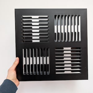 May include: A black metal vent cover with a square shape. The vent cover has a series of horizontal and vertical slots, creating a grid-like pattern. The vent is being held by a person.