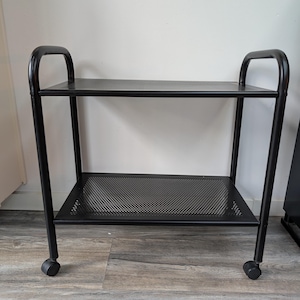 May include: Black metal rolling cart with two perforated shelves and curved handles. The cart has four small wheels for easy movement. The shelves are rectangular and the frame is made of black metal.