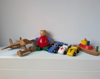 May include: A collection of wooden toys including a natural wood airplane, a bear figure with a red body and green base, a blue race car, a white ambulance with a red cross, a yellow car with a black T, and a boat with a red and yellow top.