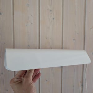 Op de afbeelding: Een witte, rechthoekige wandlamp met een gebogen rand. De lamp wordt tegen een lichtgekleurde houten muur gehouden. De lamp heeft een eenvoudig, modern ontwerp en een snoer hangt van de onderkant.