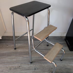 May include: A chrome-plated metal step stool featuring a black rectangular top seat and two lower steps with light brown, ribbed, non-slip surfaces. The frame is constructed from tubular steel with black protective caps on the four floor contact points.