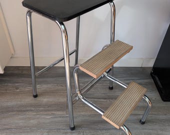 Pode incluir: Um banco de degraus em metal cromado com um assento superior retangular preto e dois degraus inferiores com superfícies estriadas antiderrapantes em castanho claro. A estrutura é de aço tubular com tampas protetoras pretas nos quatro pés.