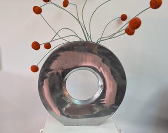 Pode incluir: Um vaso circular de metal prateado com um centro oco, apresentando uma superfície martelada e reflexiva. O vaso contém vários caules finos verde-claros cobertos por pequenas esferas de feltro redondas na cor laranja queimado.