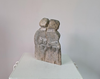 May include: A grey and beige speckled stone sculpture depicting two figures standing side by side with their arms linked. The piece features a smooth, polished texture with visible natural mineral inclusions throughout the carved stone surface.