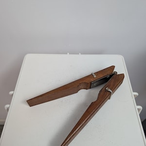 Puede incluir: Un cascanueces de madera con dos mangos largos y cónicos conectados por un mecanismo de bisagra de metal. La herramienta presenta un acabado de veta de madera marrón oscuro y herrajes de metal plateado, incluyendo pequeñas bandas metálicas.