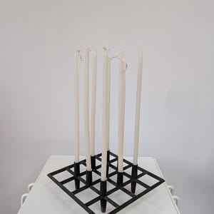 Op de afbeelding: Een zwarte metalen kandelaar met een vierkant roosterontwerp, met zes crèmekleurige taps toelopende kaarsen. De kaarsen zijn van verschillende hoogtes en hebben licht gesmolten toppen. De houder staat op een wit oppervlak.