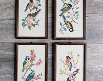 May include: A set of four rectangular framed cross-stitch embroideries on off-white fabric. Each piece features two birds perched on flowering branches with colourful thread details, including pink, blue, yellow, and green, enclosed in dark wood frames.