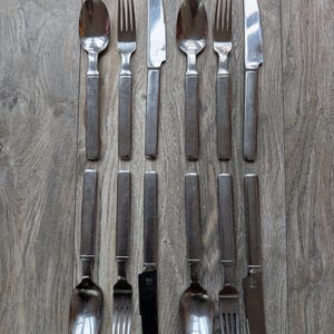 Könnte beinhalten: Ein Satz Vintage-Besteck aus Edelstahl, einschließlich Löffel, Gabeln und Messer. Das Besteck hat ein schlichtes, rechteckiges Griffdesign und eine polierte Oberfläche. Die Anordnung ist symmetrisch auf einer Holzoberfläche.