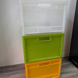 May include: A stack of three rectangular plastic storage crates. The top crate is translucent white with a grid-like structure, the middle crate is solid lime green, and the bottom crate is solid yellow. Each crate features integrated side handles.