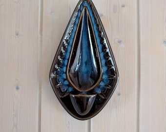 Pode incluir: Um candeeiro de parede em cerâmica em forma de lágrima com um acabamento castanho escuro brilhante e um detalhe central vidrado em azul. A peça tem uma borda texturizada e recortada em torno de um centro liso e um pequeno copo na base.