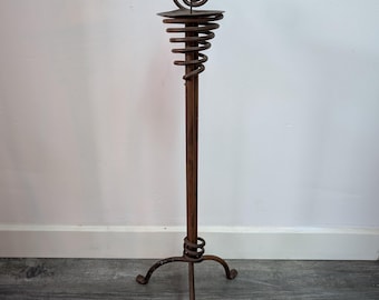 Pode incluir: Um castiçal de metal castanho-escuro alto com uma haste vertical fina apoiada numa base de tripé de três pernas. A secção superior consiste numa espiral de metal enrolada que forma uma plataforma para uma vela, rematada com um detalhe.