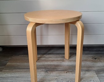 May include: A light-coloured wooden stool featuring a circular seat and three curved legs. The stool is constructed from bent plywood with a smooth, natural wood grain finish, showcasing a simple, functional design with clean lines and sturdy construction.